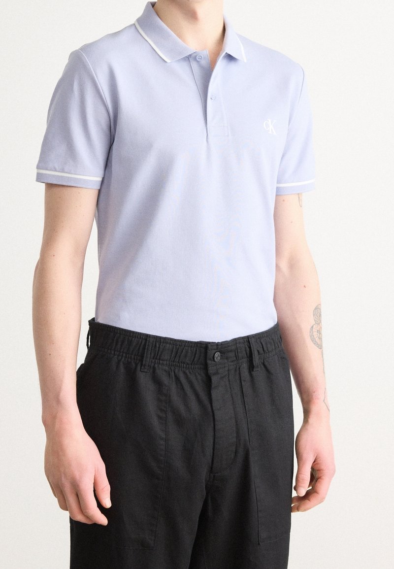 Personne portant un polo bleu clair à manches courtes avec des bordures blanches et un pantalon noir, debout devant un fond uni.