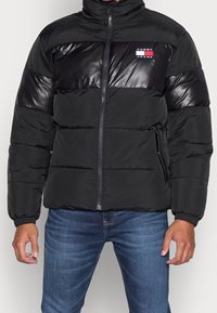 Chaqueta de plumas negra con un panel superior brillante y texturizado, cierre con cremallera, bolsillos laterales y parche de logo en el pecho. Diseño grueso e insulated.