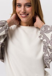 Lichtbeige top met volumineuze mouwen. De mouwen hebben een herhalend abstract bruin lijnpatroon. Glad materiaal, ronde halslijn, ontspannen pasvorm.