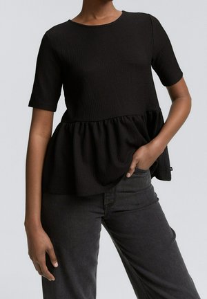 Kvinde iført en sort kortærmet peplumtop og mørkegrå jeans, med den ene hånd i lommen og den anden afslappet ved siden af.