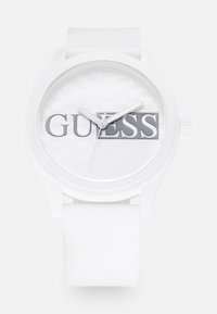 Montre en silicone blanche avec un boîtier rond, un cadran transparent arborant un motif répété 'GUESS', et des aiguilles argentées.