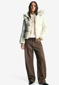 Jonge vrouw in een crème pufferjack over een beige hoodie en een paarse wijde broek, staand met één hand in haar zak tegen een witte achtergrond.