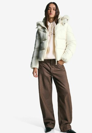 Jonge vrouw in een crème pufferjack over een beige hoodie en een paarse wijde broek, staand met één hand in haar zak tegen een witte achtergrond.