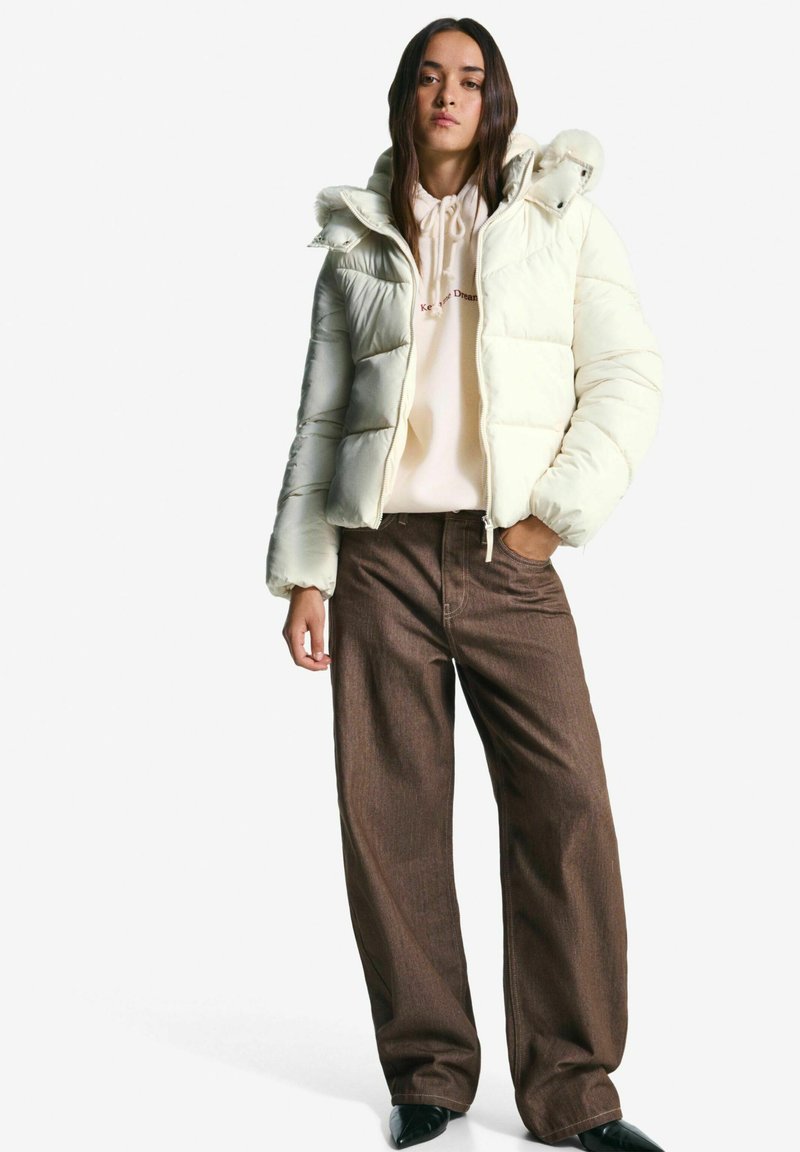Jonge vrouw in een crème pufferjack over een beige hoodie en een paarse wijde broek, staand met één hand in haar zak tegen een witte achtergrond.