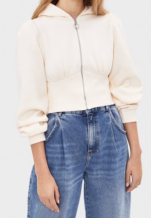 Frau trägt einen cremefarbenen, kurzen Hoodie mit Frontreißverschluss und aufgepufften Ärmeln, kombiniert mit hoch taillierter blauer Jeans.