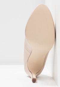 Chaussure beige à talon haut avec bout pointu et semelle lisse vue de dessous, reposant verticalement contre un mur blanc.