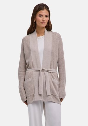 Frau trägt beige Strickjacke mit Fronttaschen und Gürtel, kombiniert mit weißem Oberteil und Hose, stehend vor einfachem Hintergrund.