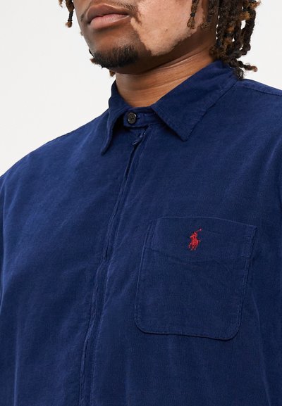 Μπλε καμβάς μπλούζα με κουμπωτό γιακά, μπροστινή τσέπη με κόκκινο λογότυπο και υφή υφάσματος. Βραχίονες κοντοί, χαλαρή εφαρμογή.