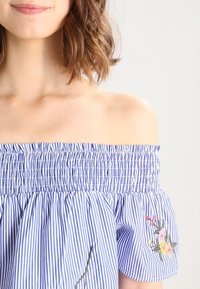 Blue and white striped off-the-shoulder top with smocked neckline, featuring floral embroidery on the left side and a textured fabric.