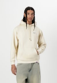Sweatshirt à capuche couleur crème avec une poche avant, présentant un design graphique sur la poitrine et des poignets côtelés. Associé à un jean en denim clair.