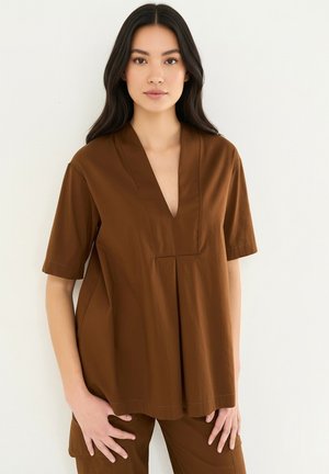 Femme aux longs cheveux foncés portant un haut marron ample à col en V et manches courtes ainsi qu’un pantalon assorti, debout devant un fond clair.