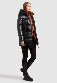 Svart vadderad jacka med huva, med glansig yta och en brun innerväst. Modellen bär en svart tröja och knähöga svarta stövlar.