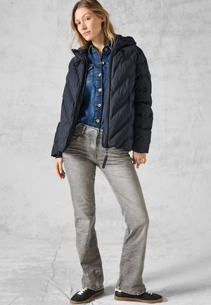 Doudoune bleu marine avec capuche, dotée d'une fermeture éclair à l'avant et de coutures diagonales. Associée à une chemise en denim et un jean gris. Baskets noires.