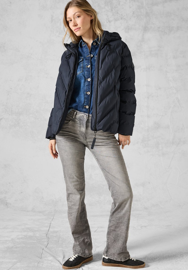 Doudoune bleu marine avec capuche, dotée d'une fermeture éclair à l'avant et de coutures diagonales. Associée à une chemise en denim et un jean gris. Baskets noires.