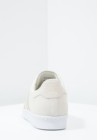 Valkoinen mokkanahkainen lenkkari, jossa on sileä pinta, pyöristetty kanta ja valkoinen kumipohja; varustettu ompeleilla ja hillityllä brändäyksellä takana.