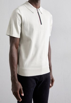 Polo de manga corta en beige claro con cuello de cremallera. Presenta una franja multicolor a lo largo de la cremallera. Textura de punto suave.