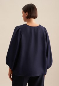 Blusa blu navy con maniche a sbuffo, scollo rotondo e tessuto morbido; le cuciture sulle spalle sono leggermente arricciate per un dettaglio di design.