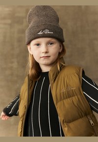 Barna dundi mellény zipzárral, fekete pulcsi felett, amelyen fehér csíkok találhatók. Barna kötött sapka "Les Deux" hímzéssel.