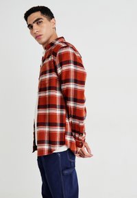 Jeune homme se tenant de côté, portant une chemise à carreaux rouge et noir sur un haut blanc avec un pantalon bleu foncé, devant un fond blanc uni.