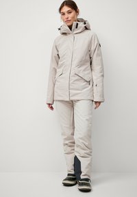 Lichtgewicht, beige waterdichte jas met verstelbare capuchon en ritsen. Gecombineerd met beige broek met donkere accenten en stevige schoenen.
