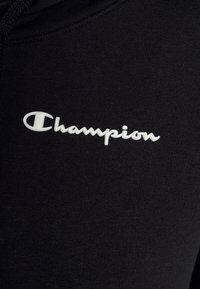 Sweatshirt noir en tissu texturé, avec un logo "Champion" blanc bien en vue brodé sur le devant.