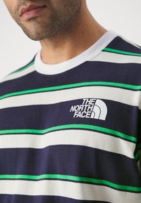 Mężczyzna w granatowej, białej i zielonej koszuli w paski z białym okrągłym dekoltem i logo "The North Face" na klatce piersiowej.