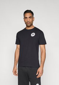 Mustast puuvillast t-särk, millel on ümmargune kaelus, lühikesed varrukad ja vasakul rinnal ringikujuline graafilise kujundusega märk.