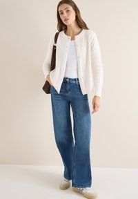 Weiße Strickjacke mit Knöpfen, getragen über einem weißen Tanktop und blauen weiten Jeans. Eine braune Umhängetasche und helle Sneakers runden das Outfit ab.