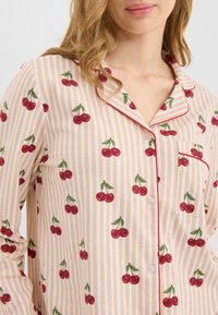 La camiseta ligera de pijama de algodón presenta rayas verticales alternadas en crema y rosa, patrones de cerezas rojas y ribete rojo a lo largo del cuello y el bolsillo.