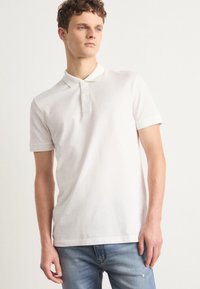 Camisa de polo blanca de algodón, con cuello, mangas cortas y un pequeño logo. Combinada con jeans de color azul claro.