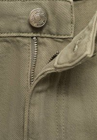 Zelená bavlnená látka s kovovým gombíkom s nápisom "DENIM AUTHENTIC." Viditeľný zips a šité švy, ktoré ukazujú hladkú textúru.