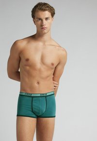 Hombre joven de pie con las manos detrás de la espalda, vistiendo bóxers de color teal con ribetes verde oscuro, contra un fondo liso.