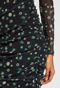 Zwarte jurk met bloemen- en paisleypatronen in groen en wit. Kenmerkt zich door geplooide details en doorschijnende lange mouwen. Zachte stoftextuur.