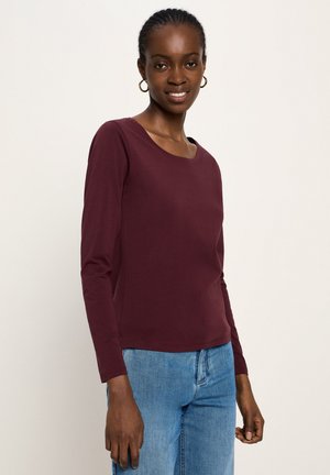 Frau trägt ein kastanienbraunes Langarmshirt und hellblaue Jeans, steht lächelnd mit goldenen Creolen vor einem einfarbigen Hintergrund.