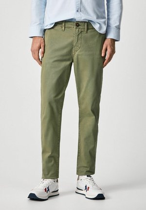 Mann trägt olivgrüne gerade geschnittene Hose, hellblaues Hemd mit Knöpfen und weiße Sneaker mit blauen und roten Akzenten.