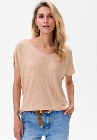 Beige T-shirt met korte mouwen en een textuur met bloemmotief, voorzien van een V-hals ontwerp. G gedragen met lichtblauwe spijkerbroek en een bruine riem.