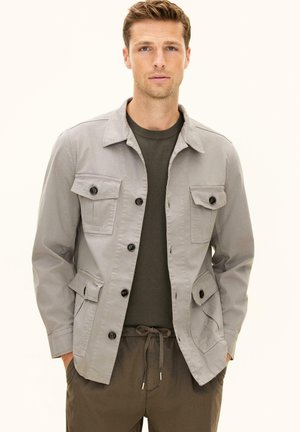 Man draagt een lichtgrijs jasje met knopen over een donkergroen overhemd en bruine broek met trekkoord, staat met handen in de zakken.