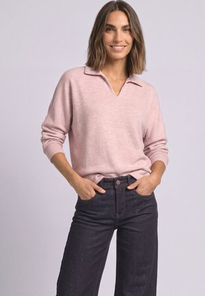 Smilende kvinde med skulderlangt hår iført en lyserød langærmet sweater og mørkeblå, højttaljede jeans med hænderne i lommerne.