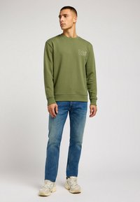 Sudadera verde oliva con cuello redondo y un pequeño logotipo bordado. Combinada con jeans azules y zapatillas de color claro.