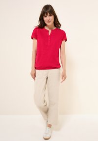 T-shirt henley à manches courtes rouge avec patte de boutonnage à trois boutons, associé à un pantalon large beige clair et des baskets blanches. Tissu lisse, coupe décontractée.