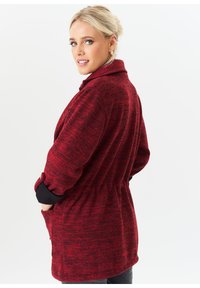 Red knit coat with a textured pattern, belted waist, roll-up sleeves, and side pockets, designed with a relaxed fit.