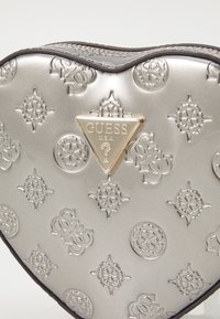Borsa a forma di cuore in ecopelle argentata con motivi in rilievo, dotata di un logo triangolare dorato "GUESS" e bordi scuri attorno alla cerniera.