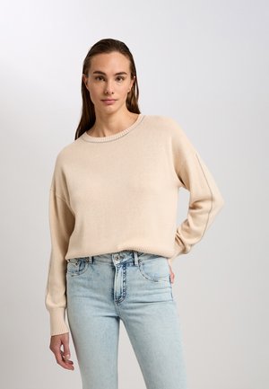 Beige Strickpullover mit rundem Halsausschnitt und überschnittenen Schultern, kombiniert mit hellblauen, hoch taillierten Jeans. Glatte Textur mit gerippten Akzenten.