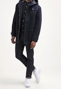 Granatowy płaszcz z kapturem z szarym podbiciem i kieszeniami zapinanymi na guziki, zestawiony z ciemnymi dżinsami i białymi trampkami, z dyskretnym wzorem na koszuli.