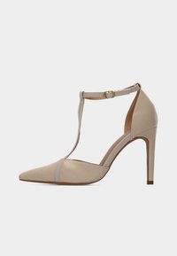 Chaussure à talon haut beige avec bout pointu, côtés ouverts et bride à la cheville. Texture lisse avec une finition brillante ; ornée d'une boucle dorée.