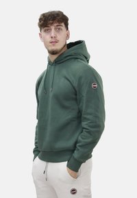 Hoodie verde con cordini regolabili, polsini a coste e una patch con logo sulla manica sinistra. Indossato con pantaloni bianchi, che mostrano un look casual.