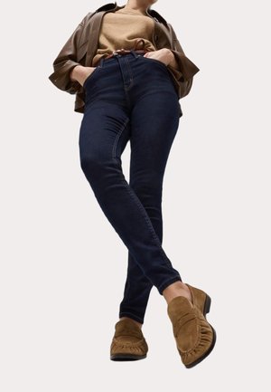 Personne portant un jean skinny bleu foncé, des mocassins en daim beige, une ceinture marron, un haut beige et une veste marron avec les mains dans les poches.