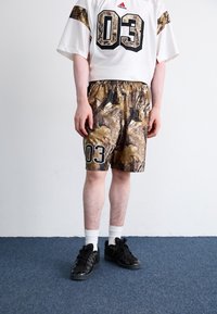 Camisola branca com o número "03" em preto e calções de camuflagem com padrões escuros. Ténis pretos com detalhes, combinados com meias brancas até ao tornozelo.