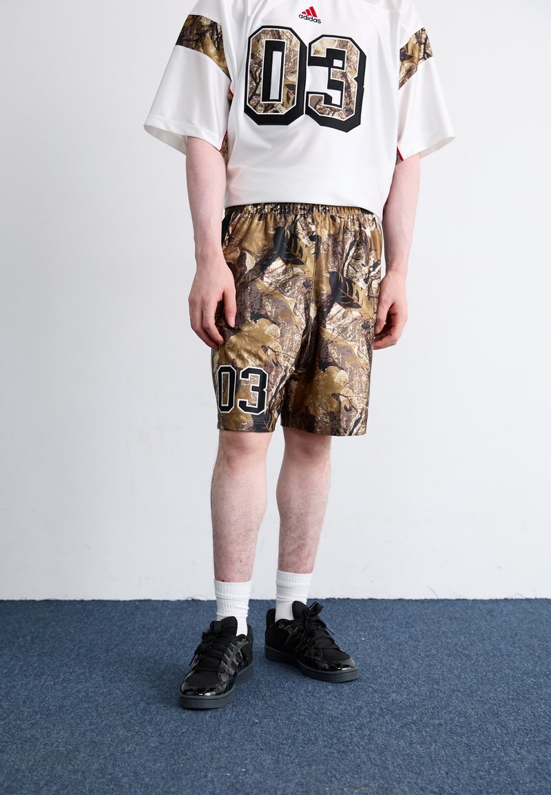 Camisola branca com o número "03" em preto e calções de camuflagem com padrões escuros. Ténis pretos com detalhes, combinados com meias brancas até ao tornozelo.