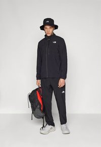 Μαύρο μπουφάν με φερμουάρ και παντελόνι, καπέλο τύπου κάδος, κρατώντας μαύρο και κόκκινο σακίδιο πλάτης. Διαθέτει λογότυπα σε όλα τα αντικείμενα, κατασκευασμένα από ελαφρύ υλικό.